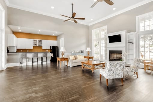 Clubhouse interior and kitchen with bar stools, wall mounted television, fireplace, wood style flooring, couch, fabric chairs, coffee table and large windows and recessed lighting