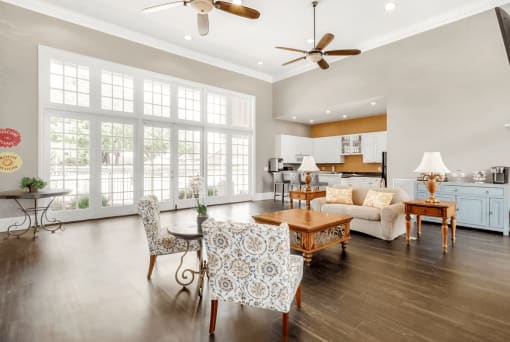 Clubhouse interior with wood style flooring, large windows and doors, ceiling fans, couch with fabric charis, coffee table and kitchen in background