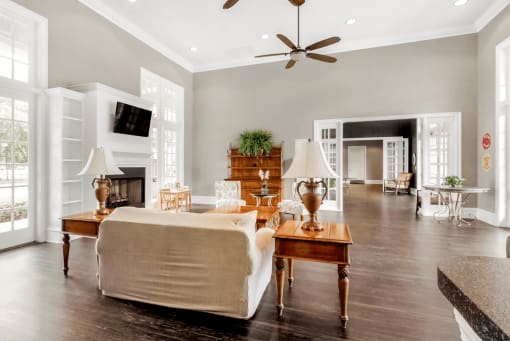 Clubhouse interior with fire place, wall mounted television, wood plank flooring, large windows, couch and chairs with coffee table and ceiling fans