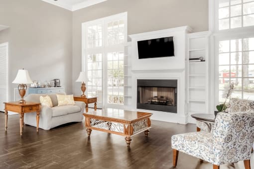 Clubhouse interior with fire place, wall mounted television, wood plank flooring, large windows, couch and chairs with coffee table