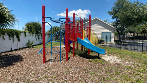 Red and blue playground set in a bed of mulch surrounded by a black fence with buildings and palm tree in the background