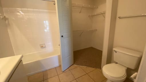 Bathroom with tile floors, toilet, shower tub combo with tile back splash, towel bar, white cabinets and view of closet with three mounted racks