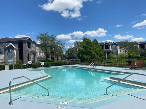 Community pool with sundeck, lounge chairs, surrounded by black metal fence with trees, lake, and building exteriors in the background