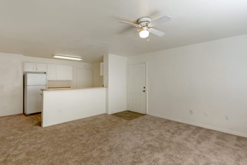 an empty living room with a ceiling fan and a kitchen in the background