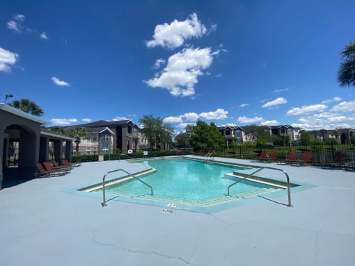 Community pool with sundeck, lounge chairs, surrounded by black metal fence with trees, lake, and building exteriors in the background
