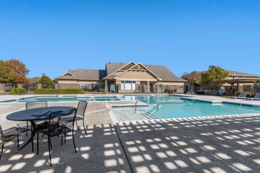 Swimming pool with large tanning deck, lounge chairs, round table and chairs with clubhouse exterior in background and native landscape