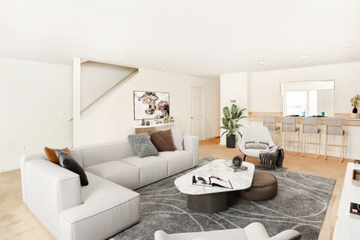 Virtually staged living room interior with grey couch, side chairs, and coffee table. Kitchen in background with grey bar stools