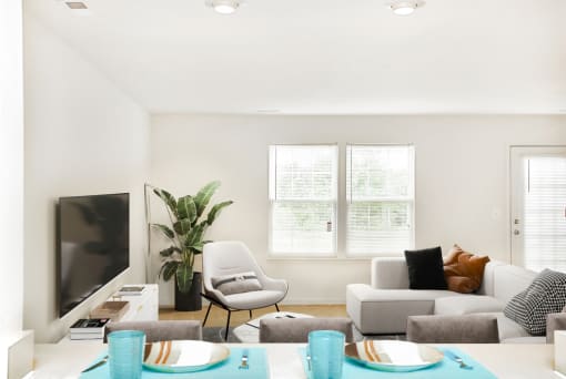 Virtually staged carpeted living room with large window next to entry door. Grey couch and accent chairs with tv in background