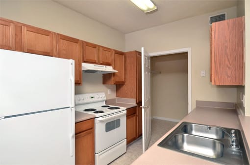 Apartment galley kitchen with brown cabinets, beige walls, and white appliances. A closet is at the far end of the room.