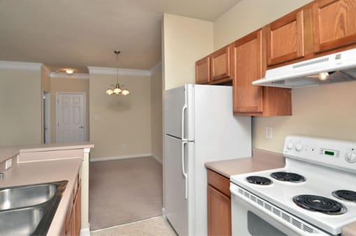 Apartment galley kitchen with brown cabinets, beige walls, and white appliances. Dining area is on the far side of the room.