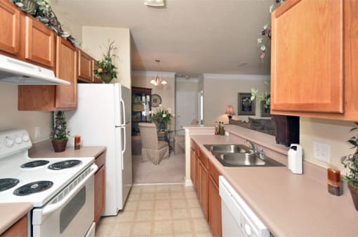 Apartment galley kitchen with brown cabinets, beige walls, and white appliances. Dining area is on the far side of the room.