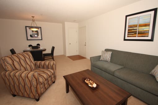 The furnished model home has hardstyle flooring in the entry by the front door with carpet in the living room and dining room. This floorplan is an open. There is a gray upholstered couch, a tan, blue, and red patterned chair, a dark wooden coffee table, a round dining table with 4 chairs and framed artwork hanging on the walls.