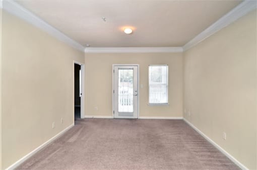 Vacant apartment living room with tan carpet and beige walls. The patio door is on the back wall of the room.