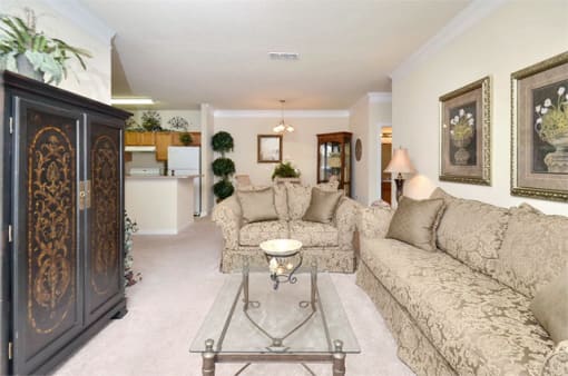 Livingroom with tan carpet, beige walls, and ceilings. Furniture throughout the room with the kitchen in the background.