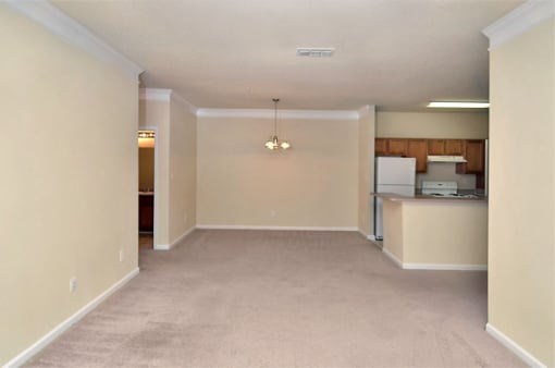 Vacant living room with tan carpet, and beige walls. the dining room and kitchen are in the background.