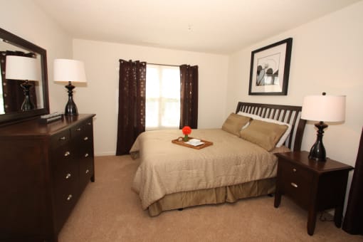 The furnished mater bedroom is carpeted throughout with a large picture window on one of the walls. Featured here is a queen size bed with tan comforter and coordinating pillows, espresso colored dresser and nightstand, and a single lamp on top of each.
