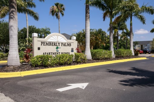 Community Entrance Sign on Parking Lot Median Surrounded by Decorative Plants with Building and Parking Lot in the Background