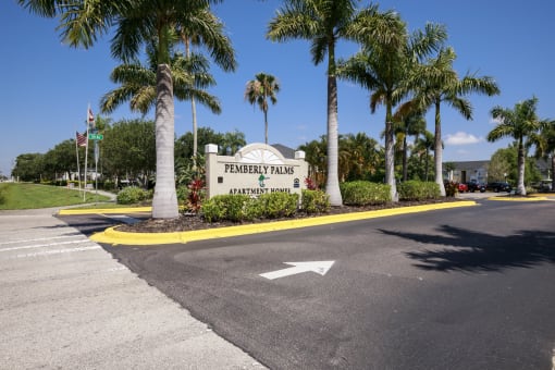 Community Entrance Sign on Parking Lot Median Surrounded by Decorative Plants with Building and Parking Lot in the Background