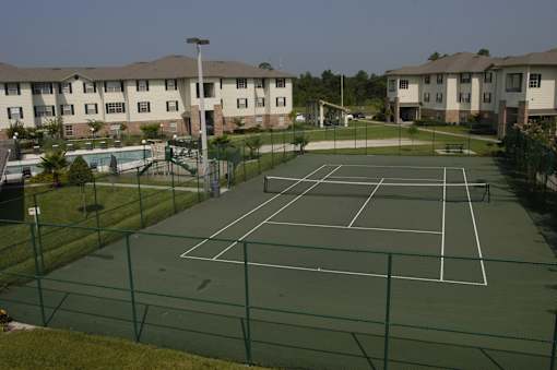 A tennis court surrounded by a green fence and apartment buildings in the background.