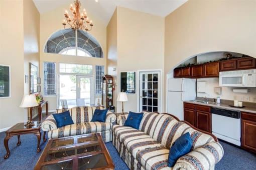 Clubhouse Seating Area with Large Windows Striped Couches with Throw Pillows Next to Coffee Table and End Table with Clubhouse Kitchen in The Background with Wood Cabinets and White Appliances