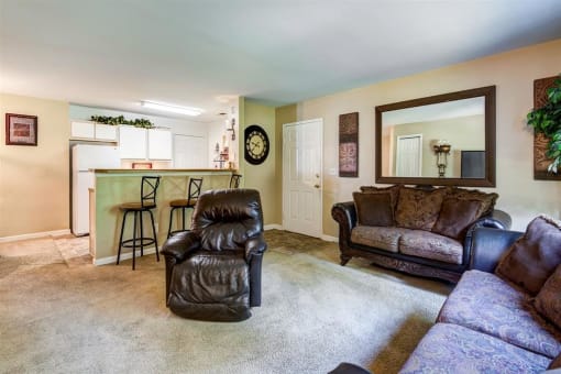 Model Living Room with a Mirror on the Wall Two Brown Couches and a Recliner and Breakfast Bar with Bar Stool and Kitchen in the Background