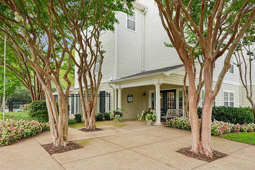 A house with a white exterior and a grey roof is surrounded by trees and flowers.