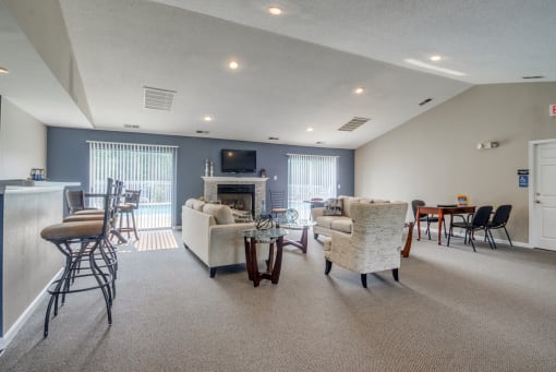 The furnished clubhouse is a large open room with a coffee bar, a full kitchen with breakfast bar, and living area with a fireplace and two sliding glass doors on either side of the fireplace. It is furnished with three bar stools, two couches and an oversized chair, two smaller tables with chairs, and a coffee table.