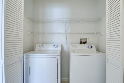 The laundry room is enclosed by two accordion, white, wooden doors. There is a washer and dryer with built-in shelving above them.