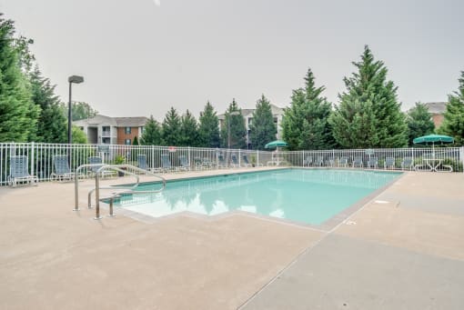 The community pool is rectangular in shape with blue and green pool furniture placed around the sundeck. There is a round picnic table with a green umbrella in the middle. The pool is placed on the back side of the Leasing Office.