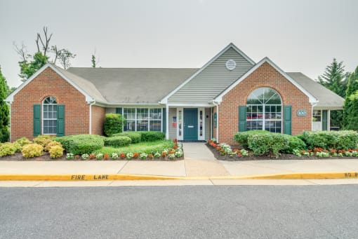 The Leasing Office building is mainly brick with a small amount of gray siding and forest green shutters and front door. There are flower beds on either side of the sidewalk leading to the front door.