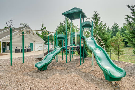 The community playground is surrounded by mulch and mature trees, and is located to the side of the clubhouse. The jungle gym is green and tan, has stairs leading to three slides and monkey bars.