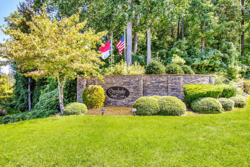Community Entrance Sign on Grass in Front of Flag Poles and Tree Line