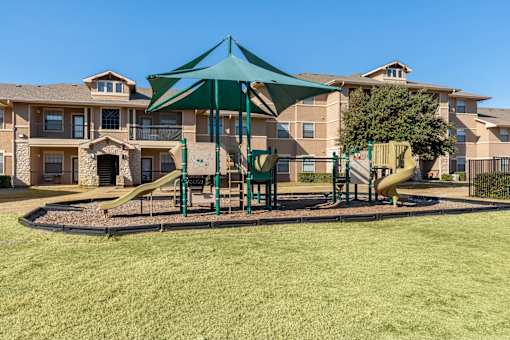 A playground with a green canopy and a yellow slide is in front of apartment buildings.