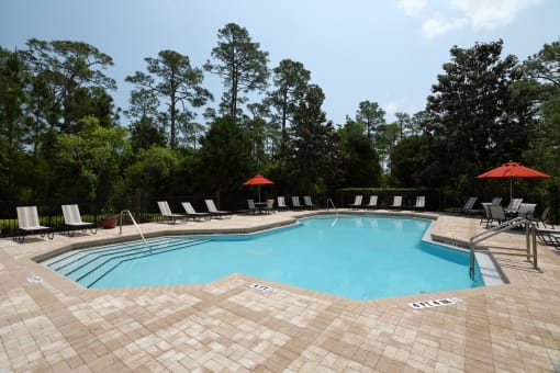The swimming pool is a large area with calm turquoise waters with a brick paver sundeck surrounding. Light lounge chairs and orange umbrellas are placed around the sundeck.