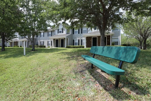 A green metal park bench sits in a grassy area of the community courtyards with trees in front of residential buildings in the background.