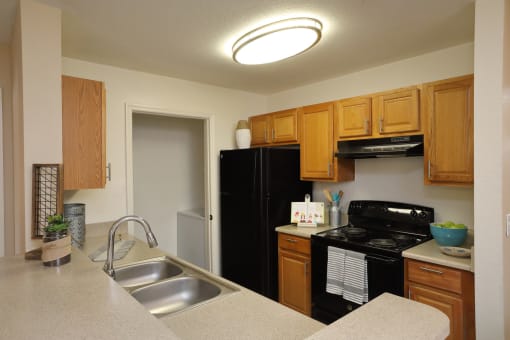 The B1 Kitchen has light oatmeal-colored countertops with light wood cabinetry. Black kitchen appliances contrast against the light paint. An open closet that at the end of the galley-style room holds the full-size washer and dryer appliances.
