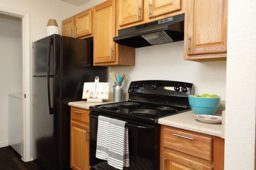 The B1 Kitchen has light oatmeal-colored countertops with light wood cabinetry. Black kitchen appliances contrast against the light paint. An open closet that at the end of the galley-style room holds the full-size washer and dryer appliances.