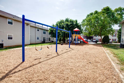 The community playground is surrounded by mulch, grass, trees and apartment buildings. It has 4 swings, and a jungle gym area with a red slide, yellow monkey bars, and stairs.