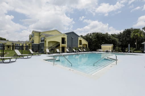 Community pool with sundeck, lounge chairs, surrounded by black metal fence with trees, and building exteriors in the background