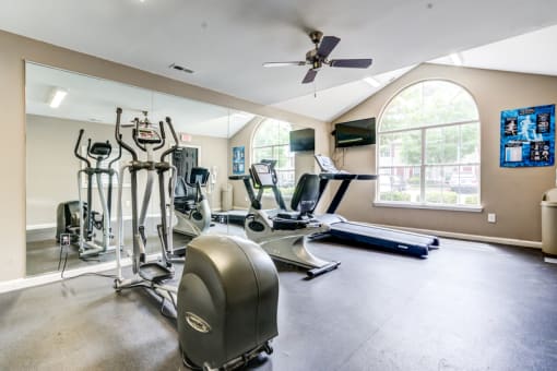 The fitness center has tan walls, rubberized flooring, a large picture window, a ceiling fan and a tv mounted on the wall. It is equipped with a treadmill, a stationary bicycle and an elliptical machine.
