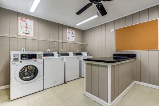The community laundry room has gray panel walls with white trim, tile flooring and a ceiling fan. It features washing machines and dryers for resident use