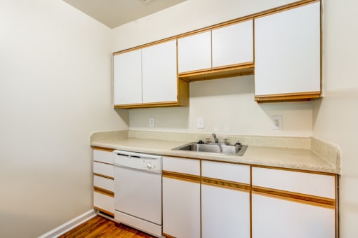 A vacant kitchen with hardwood style flooring, white walls, white cabinets with light wood trim, light countertops, a single sink and a white dishwasher