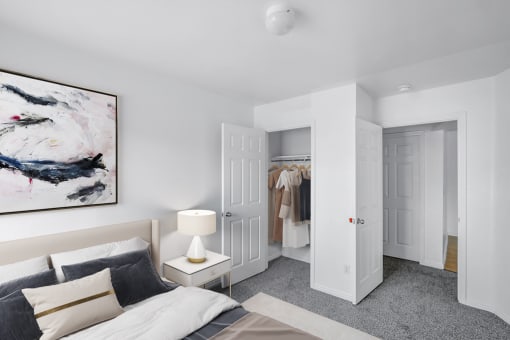 Virtually staged bedroom with bed, wall art, side table with lamp, white walls and carpet with view of closet containing hung clothesand hallway