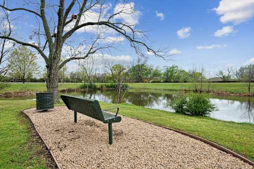 A green bench sits on a gravel path next to a tree and a body of water.