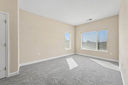 A room with a carpeted floor and two windows with blinds.