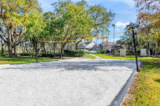 Sand Volley Ball Court Surrounded by Trees with Nearby Bench in Front of Shrubs Next to Sidewalk Leading To Parking Lot with Building Exterior in The Background