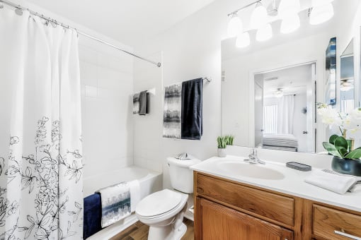 Bathroom with large mirror, countertop, toilet, and a tub and shower combo with hardwood-style flooring and modeled with towels and accessories.