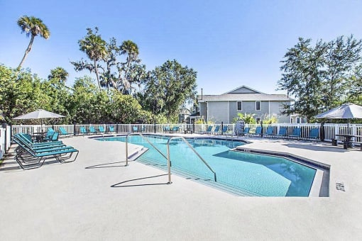 Community pool with sundeck, lounge chairs, surrounded by black metal fence with trees, lake, and building exteriors in the background