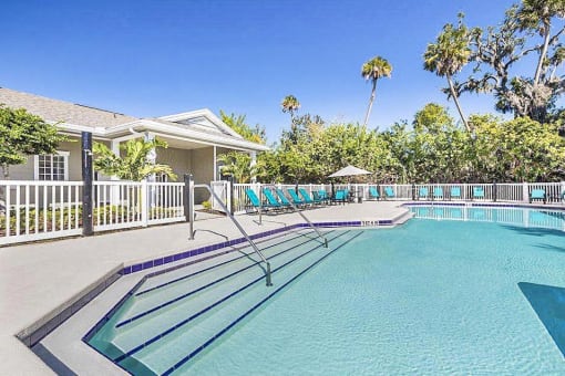 Community pool with sundeck, lounge chairs, surrounded by black metal fence with trees, lake, and building exterior in the background