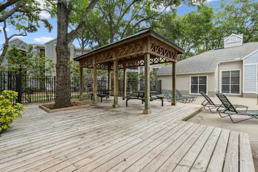 A wooden gazebo with a roof and pillars is surrounded by a deck with chairs.